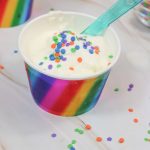 A cup with a rainbow-striped design holds vanilla ice cream made using the ice cream in a bag recipe, topped with colorful round sprinkles and served with a translucent plastic spoon.