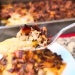 Close-up of a fork holding a delicious bite of Cheeseburger and Fries Casserole, showcasing its perfectly browned topping. In the background, a plate and baking dish are filled with more of the savory casserole.