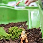 A toy tiger is carefully placed on soil with lush green moss in a vibrant container, reminiscent of a zookeeper's animal care activity. In the background, a child's hands are gently arranging its surroundings.