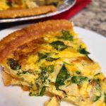A slice of spinach quiche on a white plate pairs beautifully with the flavors from a Tuna Bacon and Mushroom Quiche resting in the background on a red cloth.
