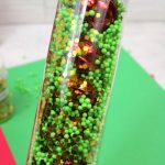 A clear bottle filled with green and gold beads, red sequins, and liquid captures the essence of winter sensory bottles. A hand with black nail polish elegantly holds it over a festive red and green background.