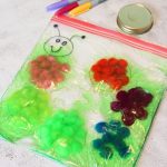 A playful caterpillar sensory bag with colorful gel compartments and a smiling face sits beside markers and a jar lid on a textured surface.