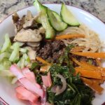A bowl featuring beef, sliced cucumber, mushrooms, pickled radish, carrots, bean sprouts, and greens on a speckled countertop.