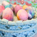 Colorful eggs dyed with shaving cream sit in a blue woven basket, with a decorated "Welcome" sign adding charm to the background.