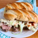 Two grilled sandwiches with leftover pork tenderloin, coleslaw, and sauce rest on a white plate. A blue and white checkered cloth sits underneath on the wooden table.