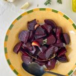 A bowl of roasted beetroot chunks, also known as roasted beets, garnished with herbs sits beside a spoon. Garlic cloves, fresh herbs, and olive oil are arranged on the table nearby.