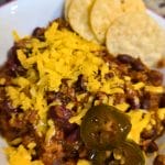 A bowl of Leftover Brisket Chili topped with shredded cheddar cheese and jalapeño slices, served with round tortilla chips on the side. Garlic bulbs visible in the background.