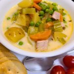 A bowl of creamy vegetable and meat soup with potatoes, carrots, peas, and celery, served with round crackers and grape tomatoes on the side.