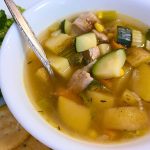 A white bowl of cozy Leftover Pork Chop Soup with diced zucchini, potatoes, and celery, served on a plate with round crackers.