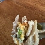A close-up of a fork holding a bite of Tuna Tetrazzini casserole with noodles, cheese, and green vegetables over a wooden table.