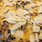 A baked casserole inspired by Southwest Tuna Noodle Casserole, topped with crumbled tortilla chips, melted cheese, black beans, and shredded meat in a metal baking pan.