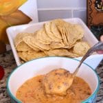A bowl of cheesy dip with ground meat is being scooped with a spoon, with a tray of tortilla chips in the background on a granite countertop.