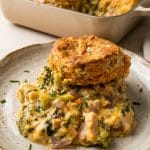 A plate with a creamy broccoli and chicken casserole topped with a golden biscuit, reminiscent of a classic tuna biscuit casserole, with a dish of the casserole in the background.